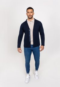 Tumesinine nööpidega kardigan, kootud tekstuuriga, koos sallikaelusega ja kahe esikotiga, kantud heleda beeži polo ja siniste teksade peal.