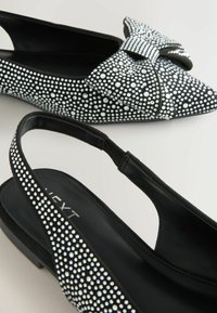 Juodos slingback bateliai su smailiais pirštais, puošti kaspinu ir dekoruoti sidabriniais rhinestones, suteikiančiais tekstūrinį ir detalų išvaizdą.