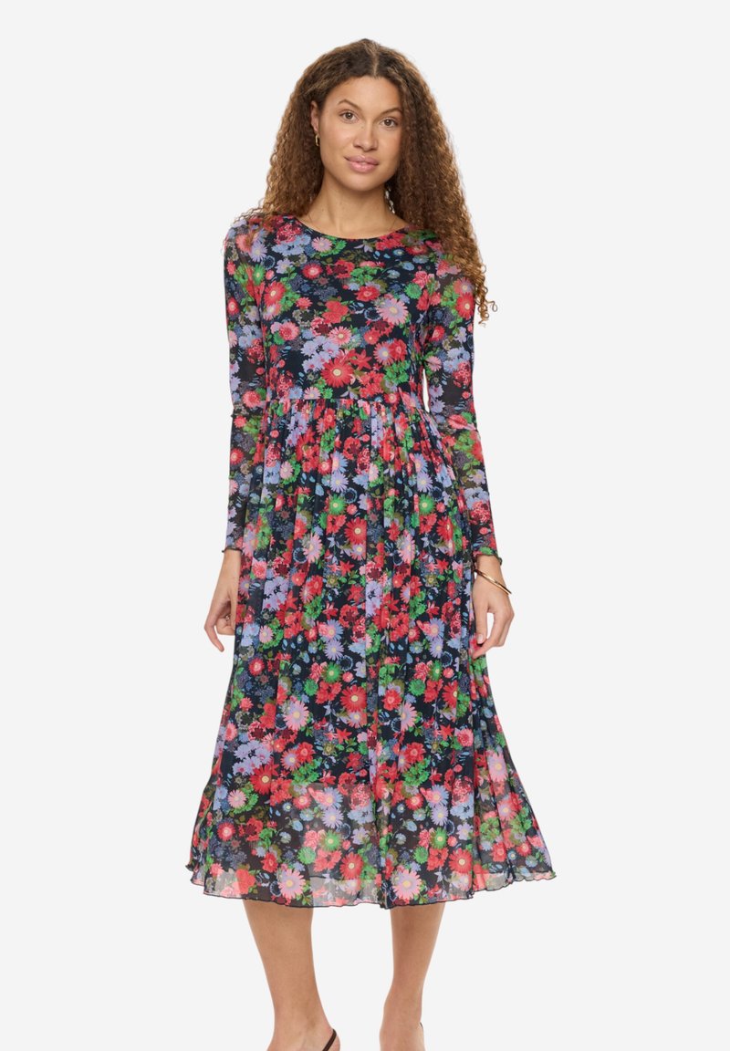Femme aux cheveux bouclés portant une robe midi à manches longues avec un motif floral multicolore, debout devant un fond clair uni.