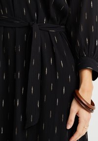 Černé šaty s texturovanou tkaninou, která má zlaté vertikální vzory. Detail s vázáním v pase a rukávy se zapínáním na gumu. Hnědý náramek na zápěstí.