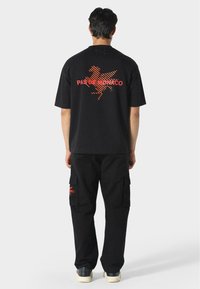 Man in een zwart oversized t-shirt en cargobroeken met een rood "Pas De Monaco" logo op de achterkant en zak, staande op een witte achtergrond.