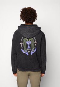 Sort hættetrøje med en falmet tekstur, der har en farverig grafik af en kalk omgivet af hænder og tekst, med lilla og grønne accenter.