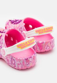 Růžové dřeváky s lesklým povrchem, se žlutým popruhem označeným "HELLO KITTY A PŘÁTELÉ" a barevnými vzory postav po celé ploše.