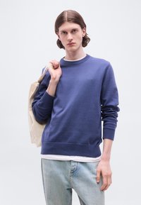 Jeune homme portant un sweat-shirt bleu BOSS et un jean clair, tenant un sac fourre-tout beige sur l'épaule, debout devant un fond uni.