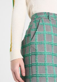 Pantalon à carreaux verts et noirs avec une finition texturée et des plis, associé à un pull crème avec des rayures vertes et jaunes sur les manches.