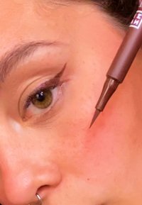 Un stylo eyeliner marron près de l'œil avec une pointe précise, appliqué le long de la paupière supérieure. La peau est claire avec des taches de rousseur et une texture visible.
