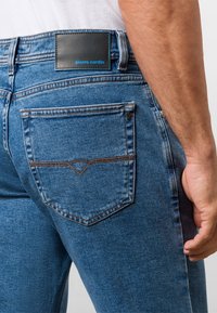 Granatowe dżinsy z tylnymi kieszeniami. Pasek w talii ma czarną skórzaną naszywkę z napisem "pierre cardin" w niebieskim kolorze. Szczegóły kontrastującego szwu.