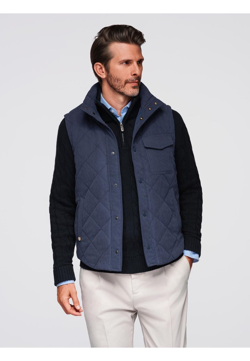 Gilet trapuntato blu navy con colletto alto, chiusura a bottoni automatici e due tasche sul petto, indossato sopra un maglione nero a maglia cable e una camicia azzurro chiaro.
