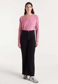 Mujer vestida con una blusa de manga larga rosa, pantalones anchos de talle alto negros y zapatillas blancas, de pie frente a un fondo liso.