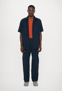 Marineblaues kurzärmeliges Hemd, orange T-Shirt und marineblaue weit geschnittene Hose. Graue Sneaker. Glatter Stoff, lockere Passform, einfaches Knopfd design.