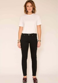 Femme aux cheveux mi-longs portant un t-shirt blanc, un jean slim noir, des mocassins bordeaux et des bracelets argentés, debout devant un fond beige.