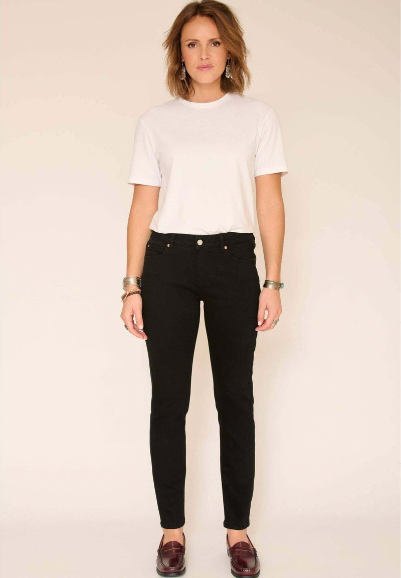 Femme aux cheveux mi-longs portant un t-shirt blanc, un jean slim noir, des mocassins bordeaux et des bracelets argentés, debout devant un fond beige.