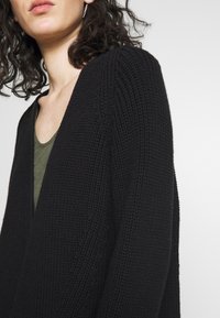 Svart stickad cardigan med V-halsdesign och texturerat tyg, i kombination med en olivgrön topp under. Har långa ärmar.