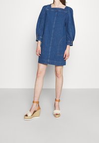 Abito in denim con scollo quadrato e maniche lunghe a sbuffo. Include due tasche laterali, accentuate da cuciture visibili. Indossato con zeppe in espadrille.