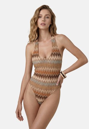 Donna che indossa un costume da bagno senza maniche a collo ad anello con motivo a zigzag in toni terrosi, con capelli mossi e braccialetti a un polso.