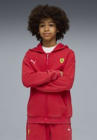 Rote Kapuzenjacke aus weichem Stoff mit einem Reißverschluss, ausgestattet mit einer Kapuze, Kängurutaschen und einem gelben Ferrari-Logo auf der Brust. Weißes Puma-Logo am Ärmel.