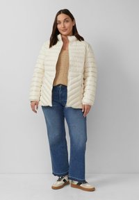 Cremfarbener Puffer-Jacke mit gestepptem Muster und hohem Kragen, kombiniert mit einem beigen Strickpullover und blauen Jeans. Klobige Sneakers runden den Look ab.