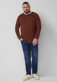 Braunes Langarmshirt mit weißem Rundhalsausschnitt, getragen zu blauen Jeans und cremefarbenen Sneakern. Das Shirt hat eine lockere Passform und eine glatte Textur.