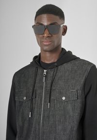 Chaleco gris oscuro con cremallera y dos bolsillos en el pecho, llevado sobre una sudadera con capucha negra. Modelo con gafas de sol negras elegantes.