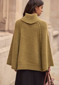 Knit poncho in olive green featuring a ribbed turtleneck, textured sleeves, and a relaxed fit, paired with a brown handbag.