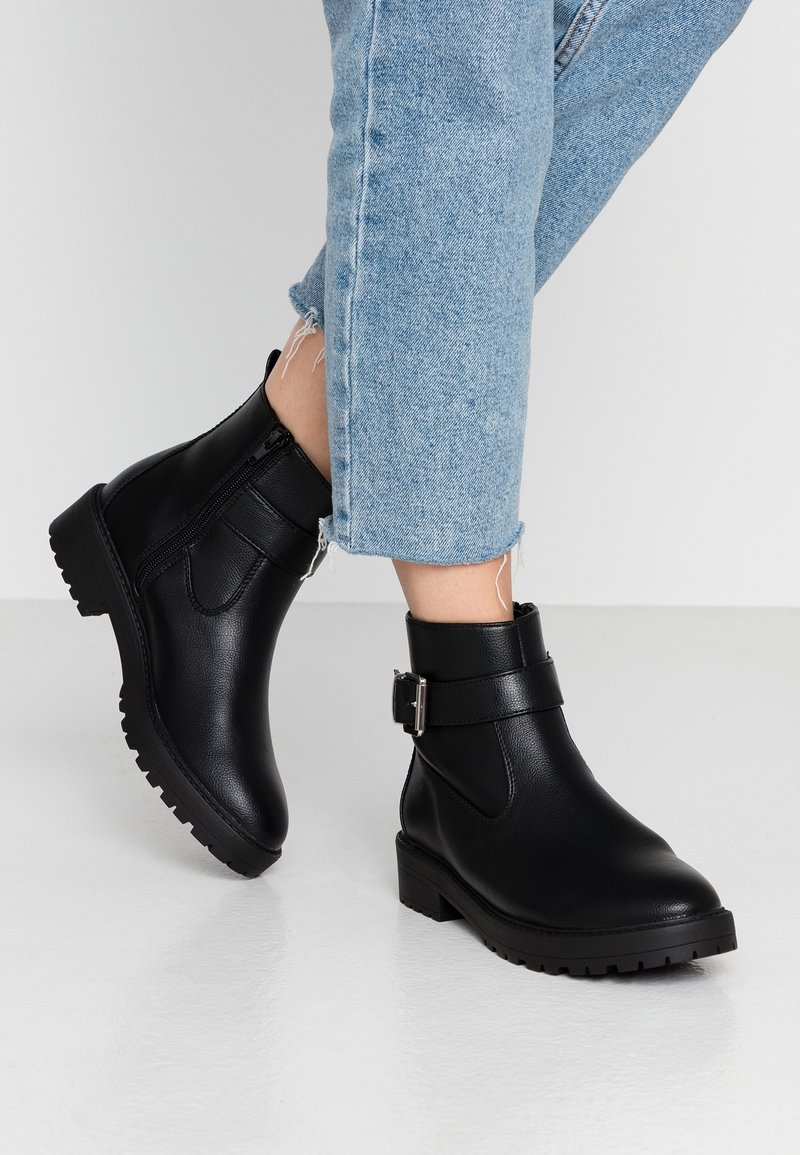 Bottes basses en cuir noir avec fermeture éclair latérale, détail de sangle à boucle, portées avec un jean bleu clair retroussé sur fond uni.