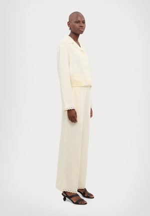 Crèmekleurige linnen tweedelige set met een geknoopte blouse met kraag en decoratieve stiksels, en wijde broek met geschulpte zoom, gecombineerd met zwarte sandaaltjes.