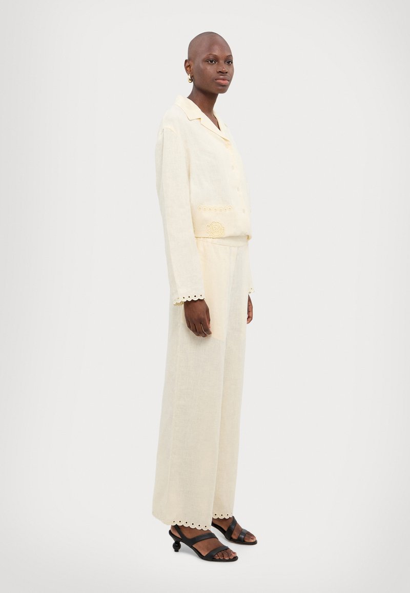 Ensemble deux pièces en lin crème comprenant une chemise à col avec couture décorative et un pantalon évasé avec des ourlets festonnés, accompagné de sandales noires.