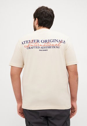Beige Baumwoll-T-Shirt mit kurzen Ärmeln. Auf der Rückseite befindet sich großer Drucktext in Marineblau und Rot. Einfaches Design, lockere Passform.