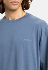 Ung person iført en blå Quiksilver t-shirt med broderet logo på brystet, som viser nakken, skulderen og dele af det krøllede hår.