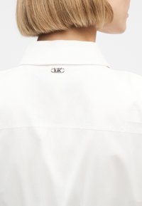 Ξανθό άτομο που φοράει λευκό πουκάμισο με γιακά και μικρό μεταλλικό λογότυπο MK στο πίσω μέρος του γιακά, στραμμένο πλαγίως με το κεφάλι γυρισμένο.