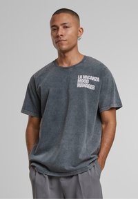 Krótki rękaw t-shirt w kolorze węgla z luźnym krojem, z białym napisem "LA VACANZA MOOD MANAGER" na lewej stronie klatki piersiowej. Miękka tekstura materiału.