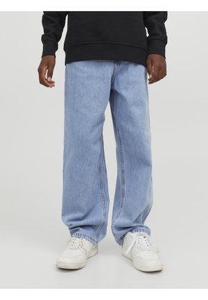 Person iført lyseblå jeans med vide ben, hvide sneakers og en sort sweatshirt, stående foran en ensfarvet baggrund.