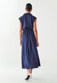 Abito maxi senza maniche di colore blu navy con colletto, vita cintrata e silhouette fluida. Indossato con mule bianchi con tacco alto.