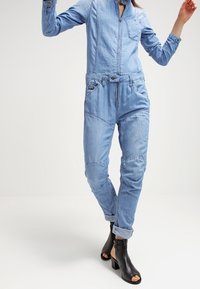 Jeansoverall i ljusblått, med dragkedja framtill, bröstficka och elastisk midja. Uppvikta ärmar och avsmalnande ben ökar komforten.