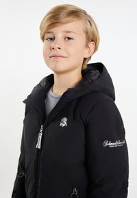 Svart pufferjacka med huva, silverdragkedja och logotyp på bröstet. Har två sidofickor och en slät yta.