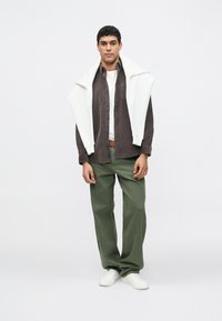 Άνδρας που φοράει σκούρο πουκάμισο με κουμπιά πάνω από λευκό μπλουζάκι, χακί παντελόνι και λευκά αθλητικά παπούτσια, με ένα λευκό φούτερ περασμένο στους ώμους.