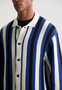 Neuletakki, jossa vuorottelevat siniset, mustat ja valkoiset raidat. Tekstuuriinen kangas, klassinen kaulus ja mustat napit.