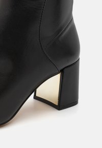 Bottines en cuir noir avec une forme angulaire, présentant un accent doré métallisé sur le talon carré. Texture lisse avec un minimum de détails de couture.