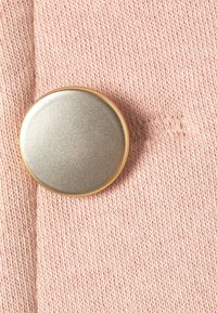 Bouton en métal rond avec une légère brillance, entouré d'un tissu tricoté rose pâle, présentant une texture subtile et une couture fine.