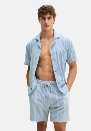Męski model ubrany w jasnoniebieską koszulę na guziki z krótkim rękawem oraz pasujące szorty na troczki z pionowymi paskami, stojący z rękami w kieszeniach.