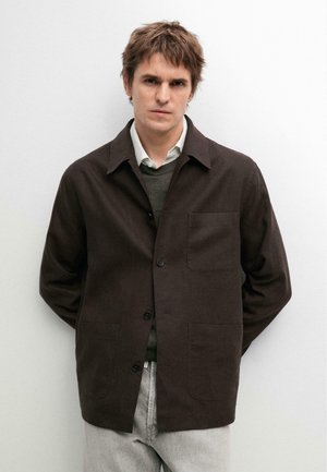 Mann mit zerzaustem braunem Haar, der eine dunkelbraune, geknöpfte Jacke über einem grünen Pullover und einem weißen Hemd mit Kragen trägt, steht vor einer weißen Wand.