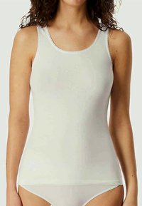 Žena v bezrukávovém bílém triku a odpovídajícím bílém spodním prádle, zobrazená od ramen po boky na jednobarevném pozadí.