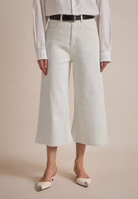 Weiße, weit geschnittene Denim-Culottes mit hoher Taille, die über Gürtelschlaufen und eine rohere Kante verfügen. Kombiniert mit weißen Slingback-Schuhen mit spitzer Zehenpartie.