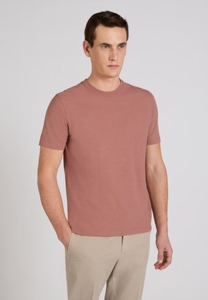 Mężczyzna w różowej koszulce z krótkim rękawem i beżowych spodniach, stojący z jedną ręką w kieszeni na jednolitym jasnym tle.