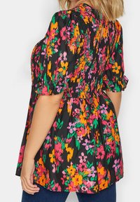 Donna che indossa una blusa floreale nera con fiori arancioni, rosa e verdi, caratterizzata da dettagli arricciati e maniche corte a sbuffo con bordi a volantino.