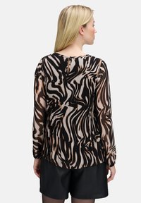 Blouse translucide avec un motif ondulé noir et marron. Présente des manches longues et une texture plissée. Encolure arrondie avec une fermeture à bouton au dos.
