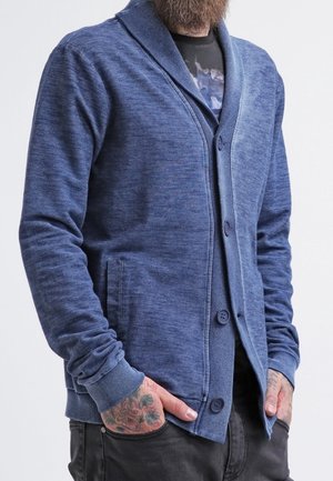 Mand iført en blå knapcardigan over en sort t-shirt med grafik, med tatoverede hænder i lommerne og mørke bukser.
