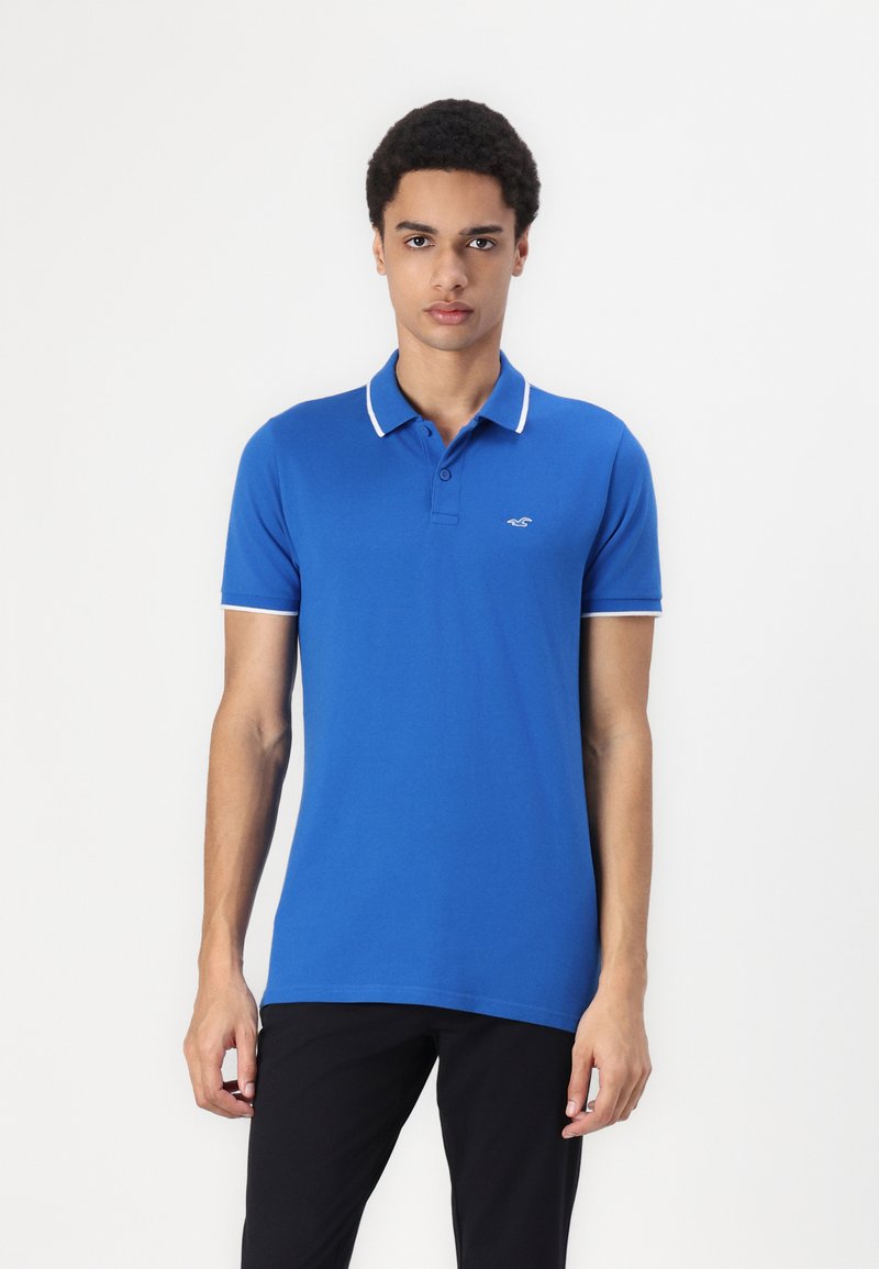 Polo bleu avec bordure blanche au col et logo sur la poitrine. Manches courtes, coupe régulière, fabriqué en tissu coton lisse. Design classique.