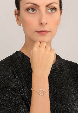Zartes Roségold-Armband mit einem runden Anhänger mit strukturierter Oberfläche, getragen an einer Hand über einem schwarzen strukturierten Oberteil.