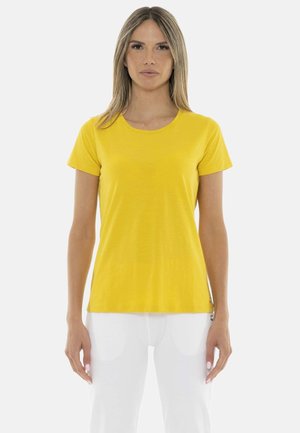 Vrouw staat met het gezicht naar voren, draagt een effen geel T-shirt met korte mouwen en een witte broek tegen een witte achtergrond.